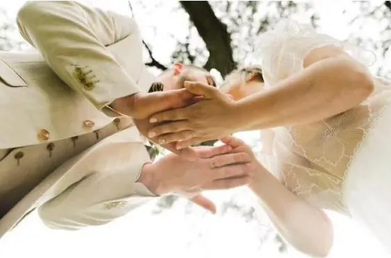 Les mains des mariés réunies, photographiées en contre-plongée sous les arbres du parc